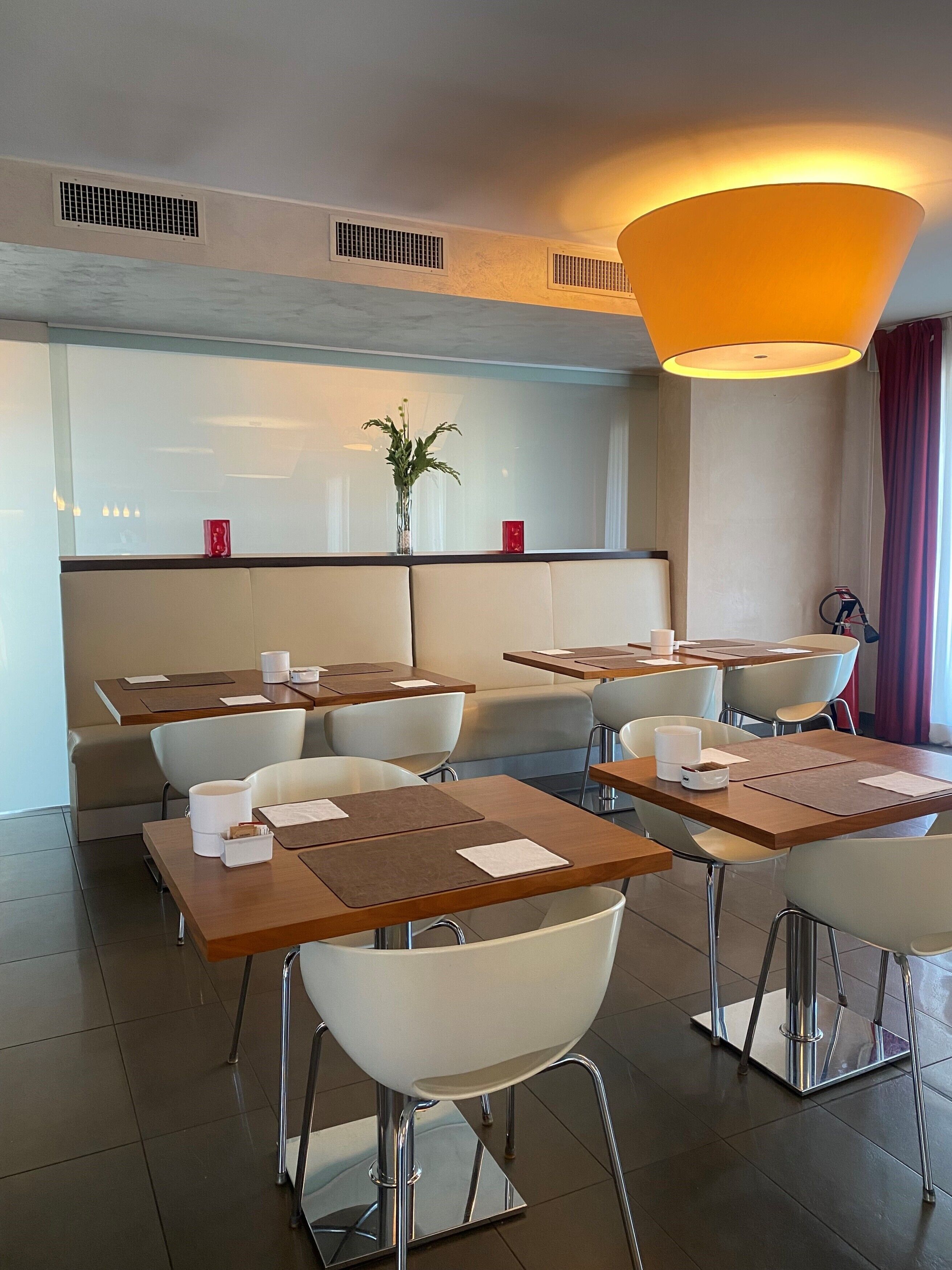 A modern dining area with tables and chairs, a central plant, and a large overhead light fixture.