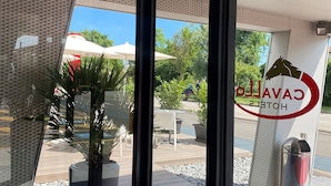 Entrada de un hotel con puerta de cristal, plantas en macetas, mesas al aire libre y vistas despejadas del entorno.
