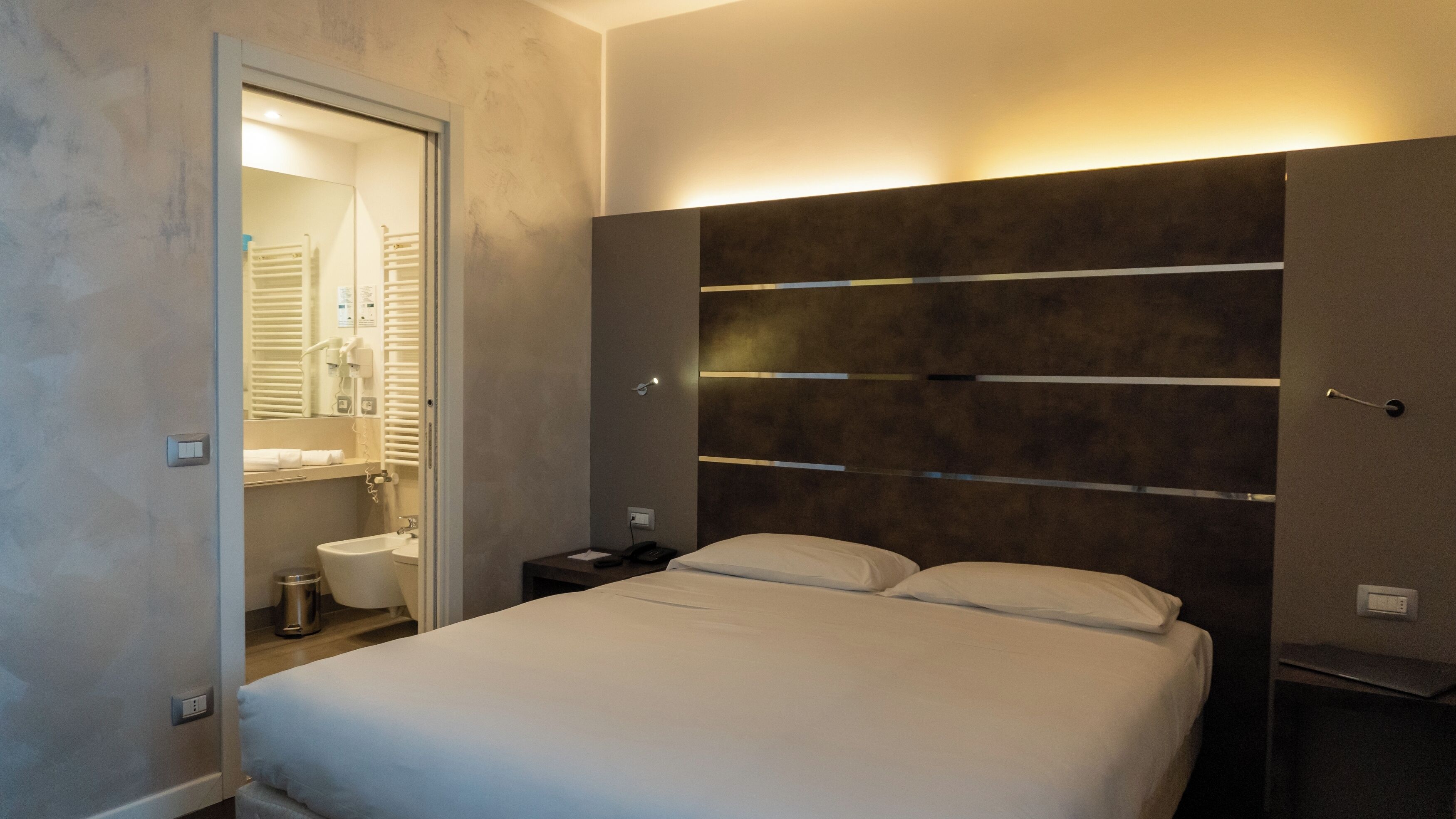 Une chambre d’hôtel moderne équipée d’un lit, d’une table de chevet, d’une commode, d’un miroir et d’une salle de bain visible à travers une porte ouverte.