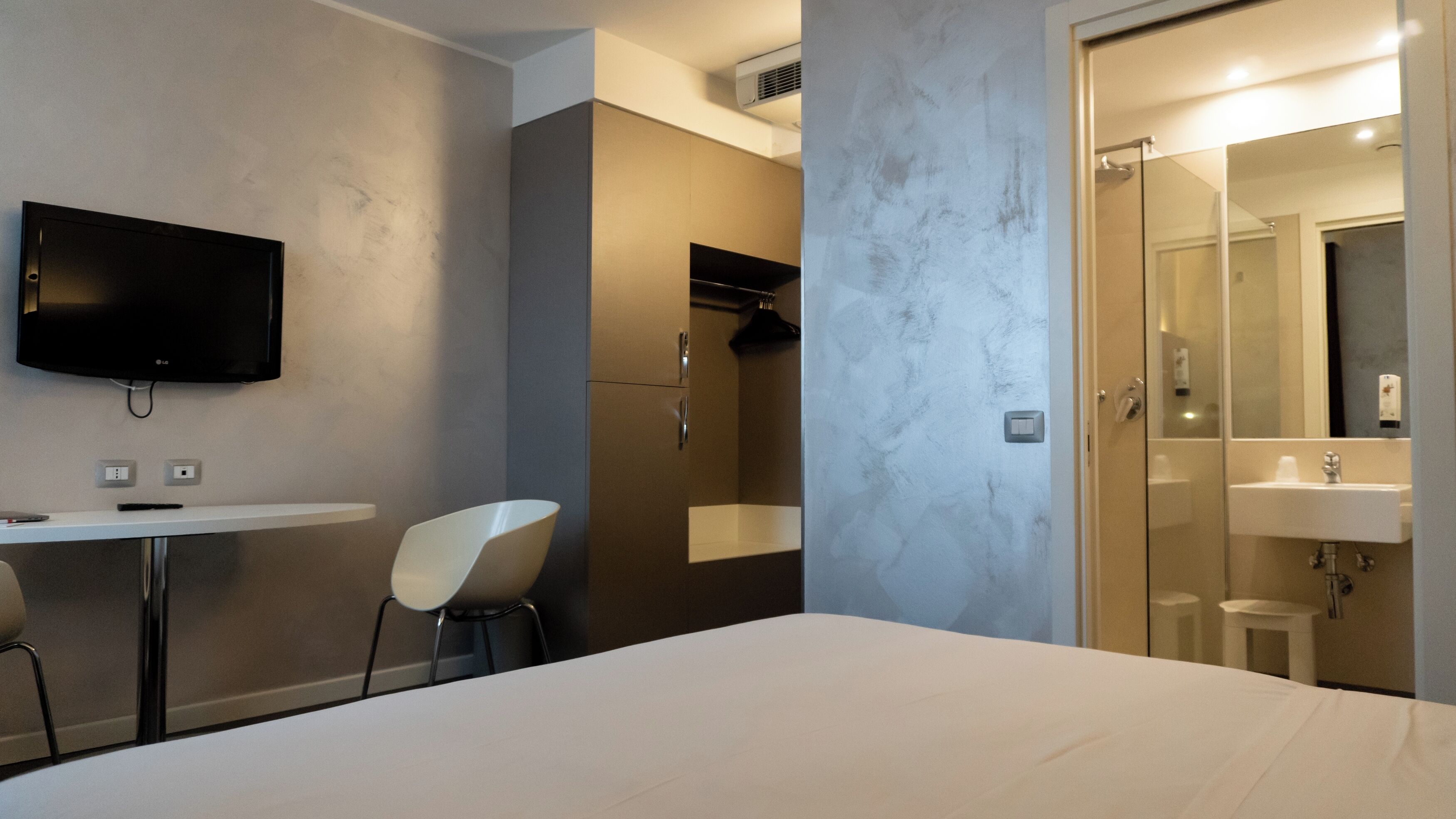 Une chambre d’hôtel moderne équipée d’un lit, d’un bureau avec une chaise, d’une télévision, d’un espace douche et d’une salle de bain avec un lavabo et un miroir.