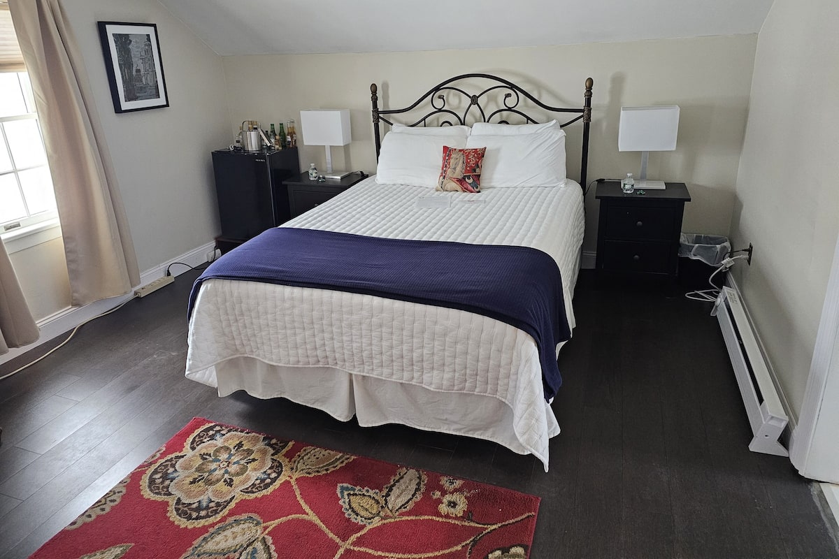 A neatly arranged bedroom with a large bed, two bedside tables, a nightstand, a radiator, and a window with curtains.