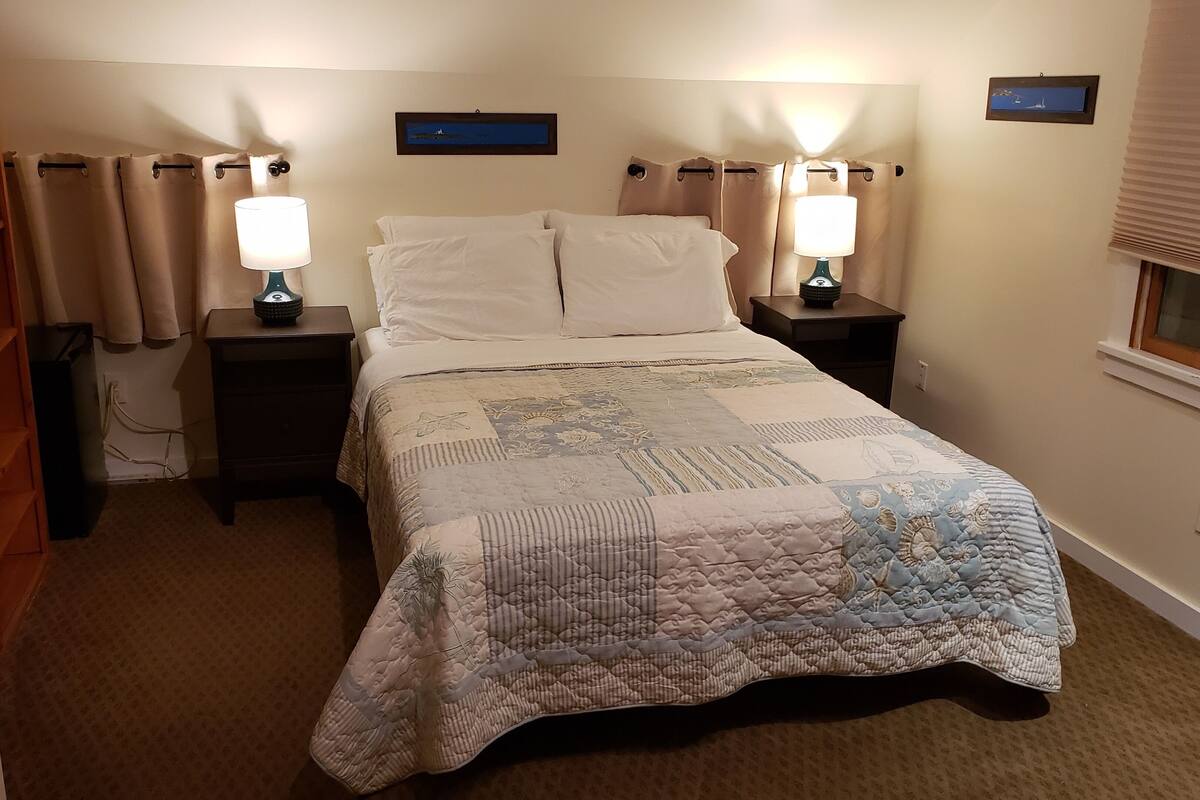 A neatly made bed with a quilt, two bedside lamps, and a window with blinds.