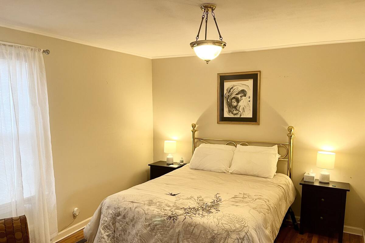 A bedroom with a bed, two bedside tables, a nightstand, a framed picture on the wall, and a hanging light fixture.