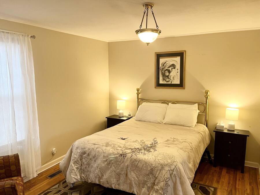 Quarto com cama, duas mesas de cabeceira, um criado-mudo e um quadro pendurado na parede, além de um lustre.