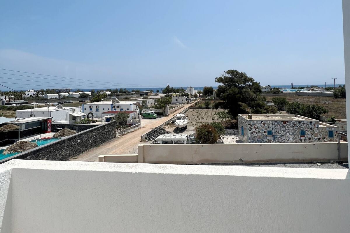 Un balcon donnant sur un quartier résidentiel, avec des maisons, une voiture verte et une étendue d’eau visible au loin.