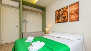 A hotel room with a bed, green bedspread, white pillows, a wooden nightstand, a lamp, a wall-mounted painting, and a wardrobe with hanging clothes. - Appartamento Ulivo in Sal (Salò)