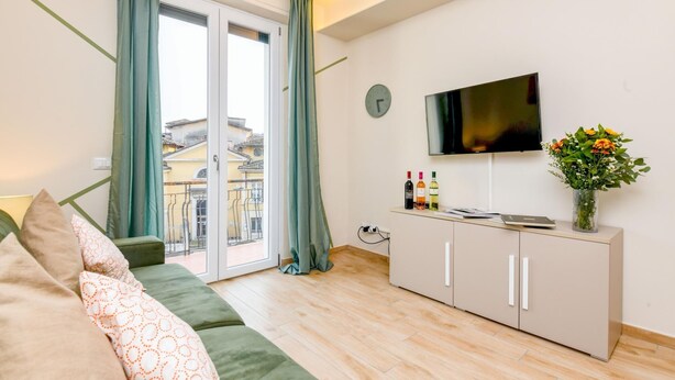A modern living room with a sofa, a flat-screen TV, a cabinet with wine bottles, and a view of buildings through a glass door. - Appartamento Ulivo in Sal (Salò)