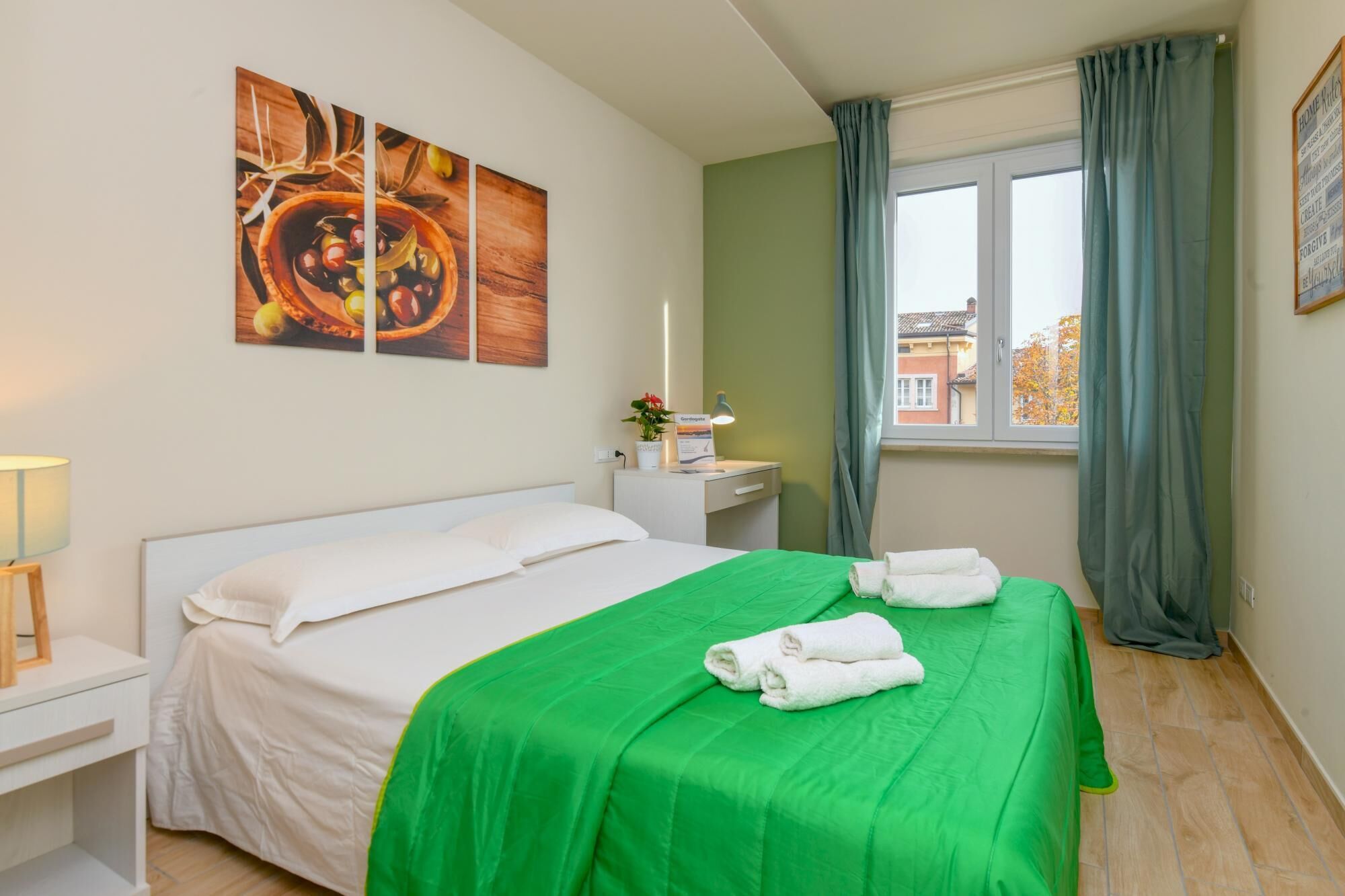 Et hotelværelse med seng, grøn dyne, hvide puder, et natbord med lampe, et vindue med gardiner og en væg med tre billeder.