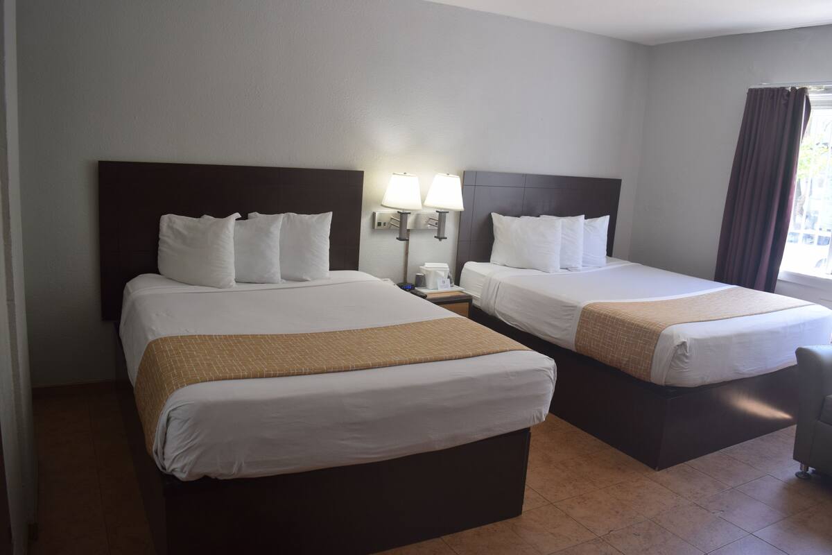 A hotel room with two beds, each with white linens and a brown throw blanket. There is a nightstand with a lamp between the beds.