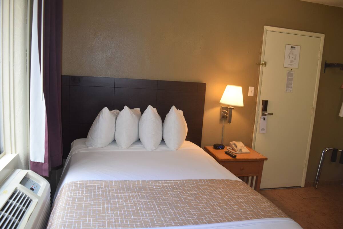 A hotel room with a bed, a nightstand with a lamp, a phone, and a door.