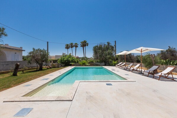 A rectangular swimming pool with a clear blue water, surrounded by a light-colored deck. There are several lounge chairs and umbrellas arranged around the pool.