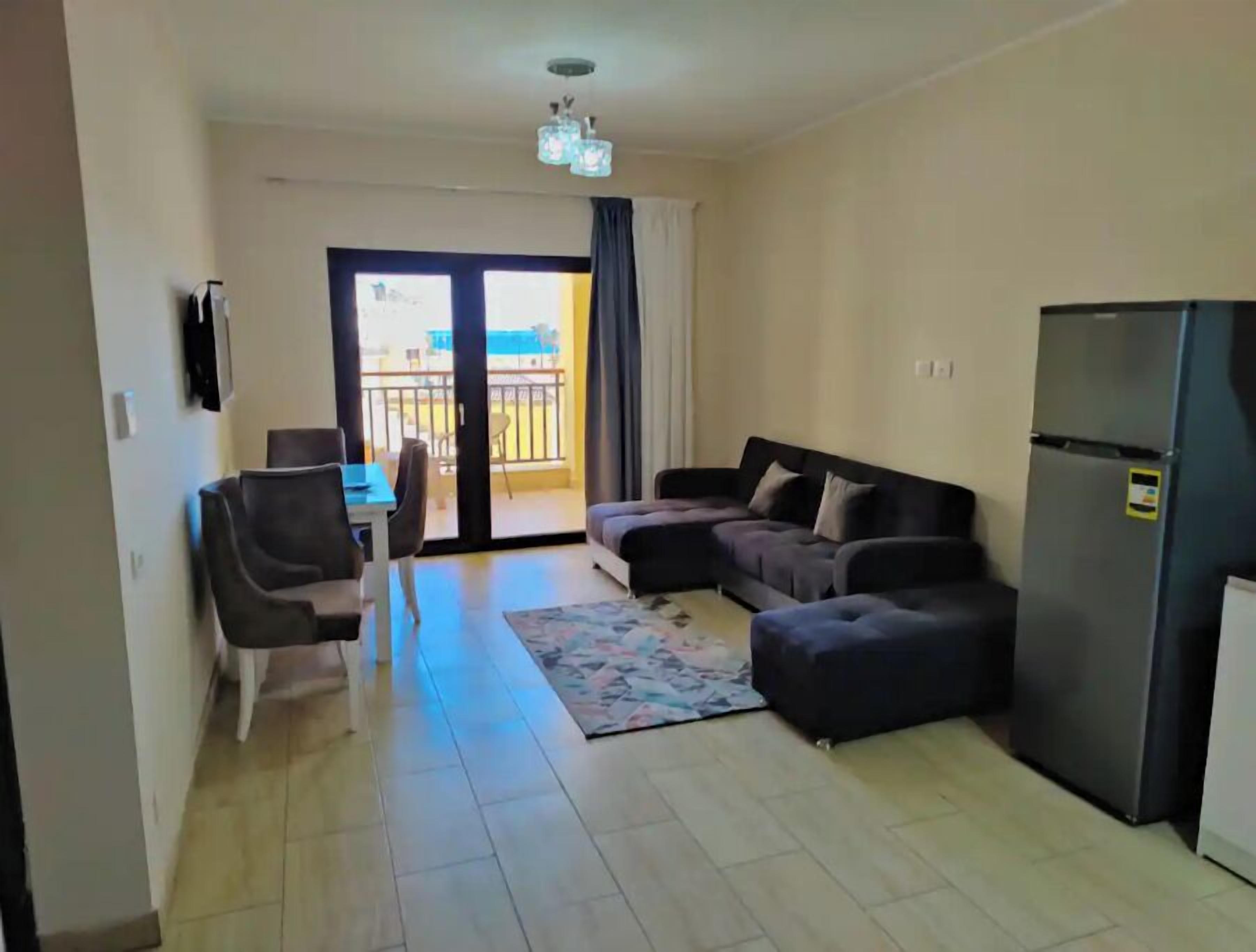 A living room with a sofa, a dining table with chairs, a refrigerator, and a view through a glass door to an outdoor balcony.