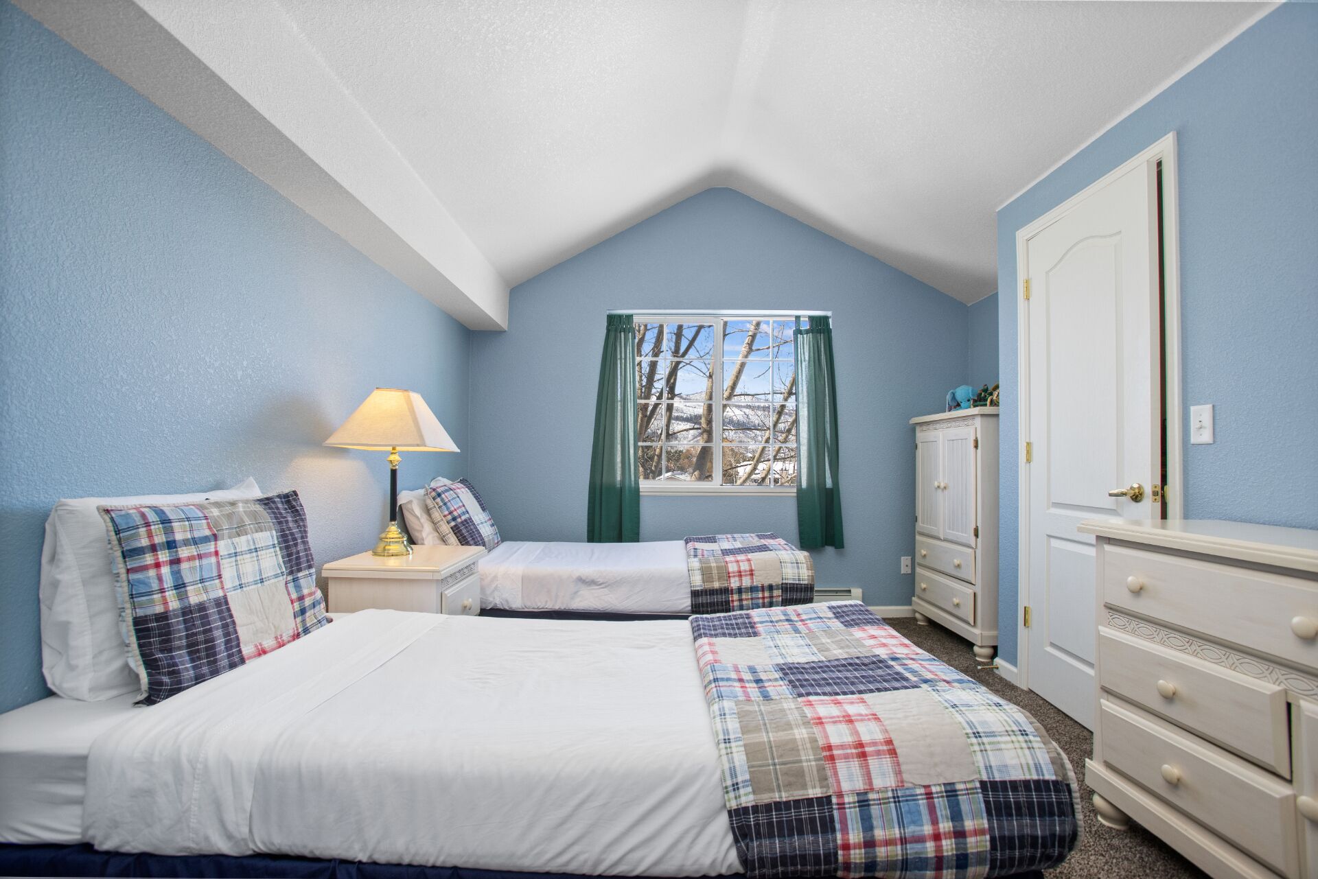 A bedroom with two beds, a nightstand with a lamp, a dresser, and a window with curtains.