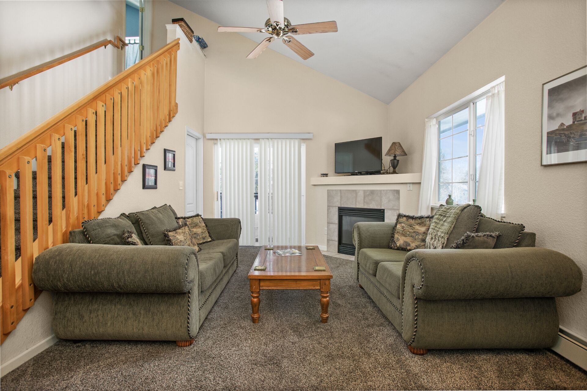 A living room with two green sofas, a wooden coffee table, a fireplace, a TV, and a staircase with wooden railings.