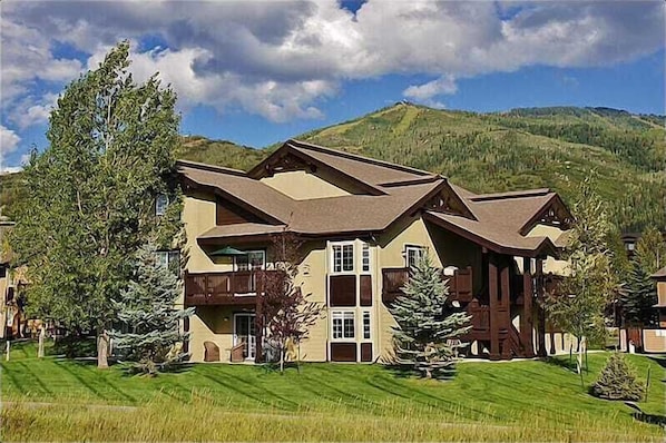A large, multi-story building with a sloped roof, surrounded by lush greenery and mountains in the background.