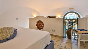 A bedroom with a bed, a decorative wall piece, a small table with a vase, and a view through a window. - The Window to the sky (Amalfi)