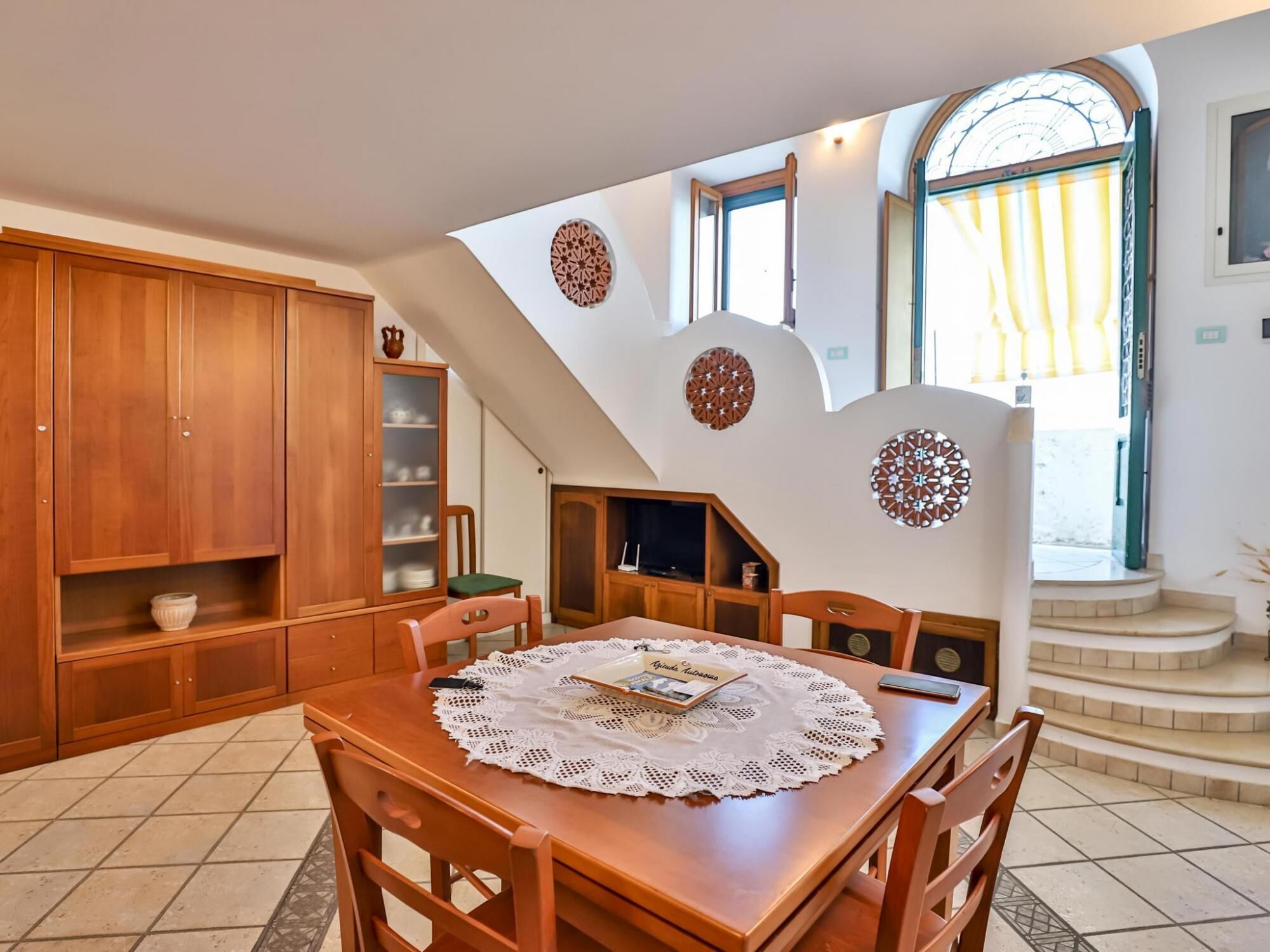 Un comedor con una mesa de madera, sillas y un camino de mesa de encaje. Hay un armario con puertas de cristal y una escalera que sube a un nivel superior.