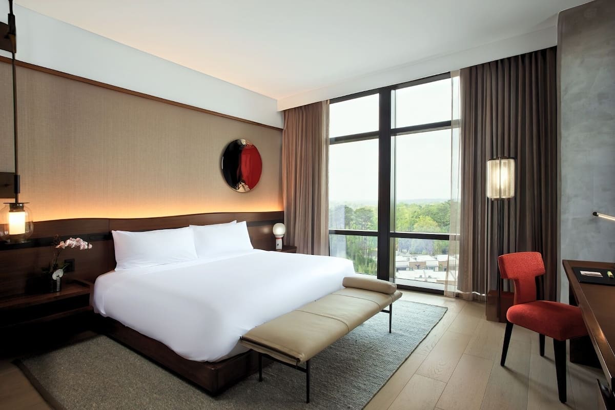 A hotel room with a large bed, a desk, a chair, and a view of the city through large windows.