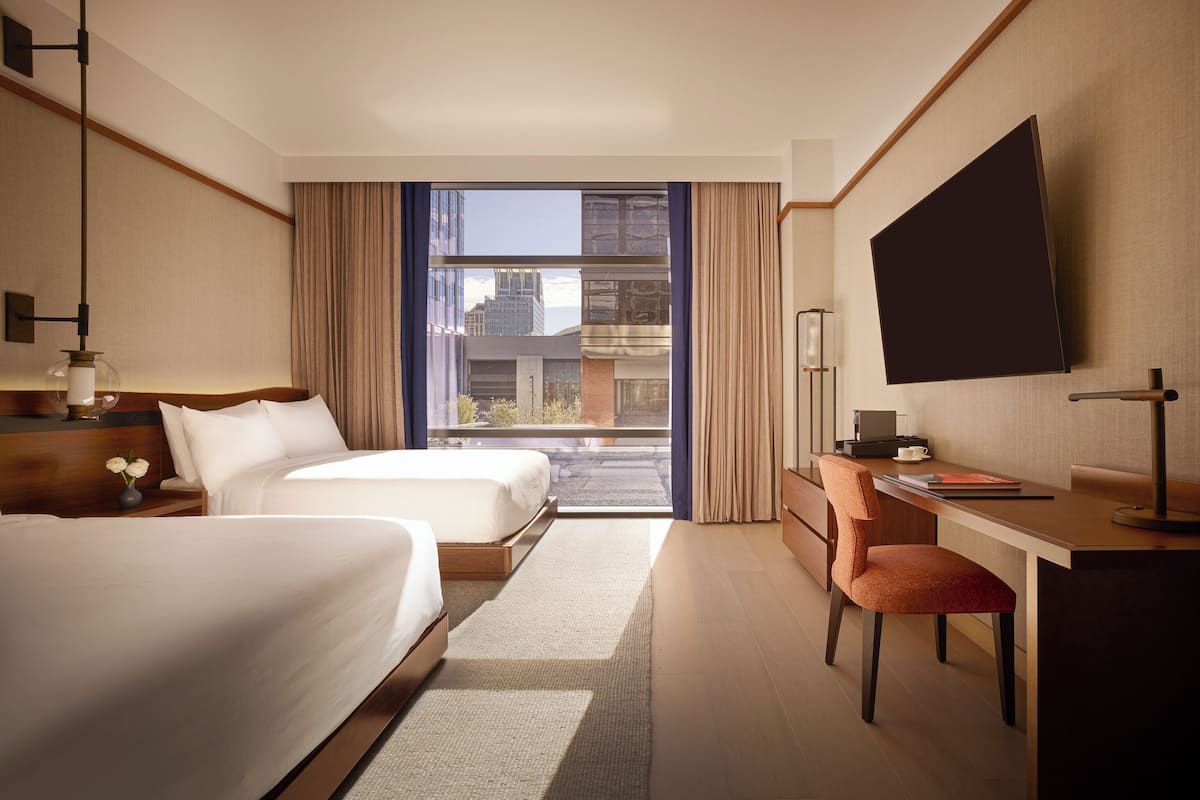 A hotel room with two beds, a desk, a chair, a TV, and a view of the cityscape.