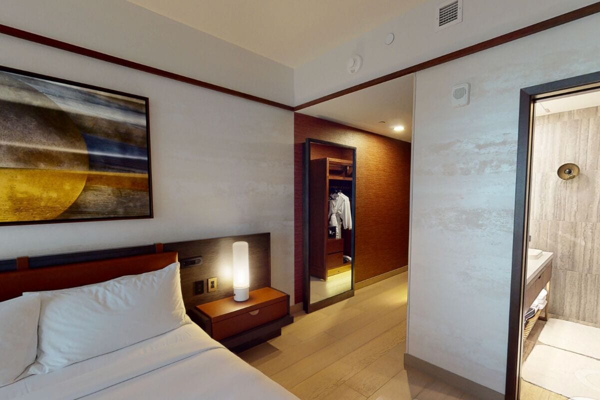 A hotel room with a bed, a nightstand with a lamp, a mirror, and a bathroom with a sink and toilet.
