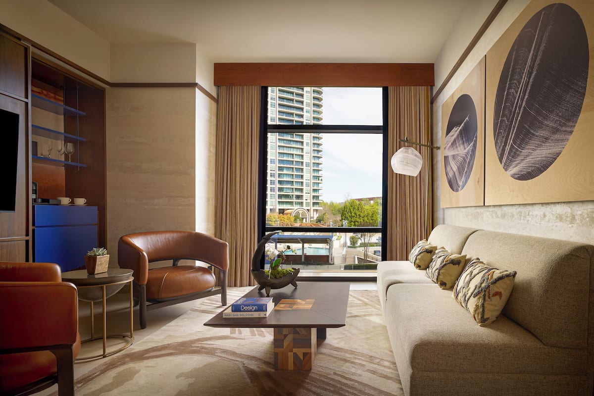 A modern hotel room with a large window, a sofa, a chair, a coffee table, and a bookshelf.
