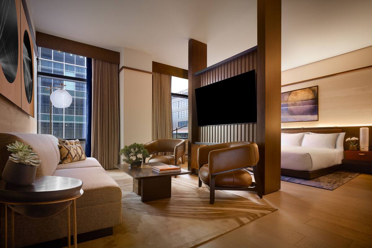 A modern hotel room with a sofa, armchair, coffee table, and a flat-screen TV.
