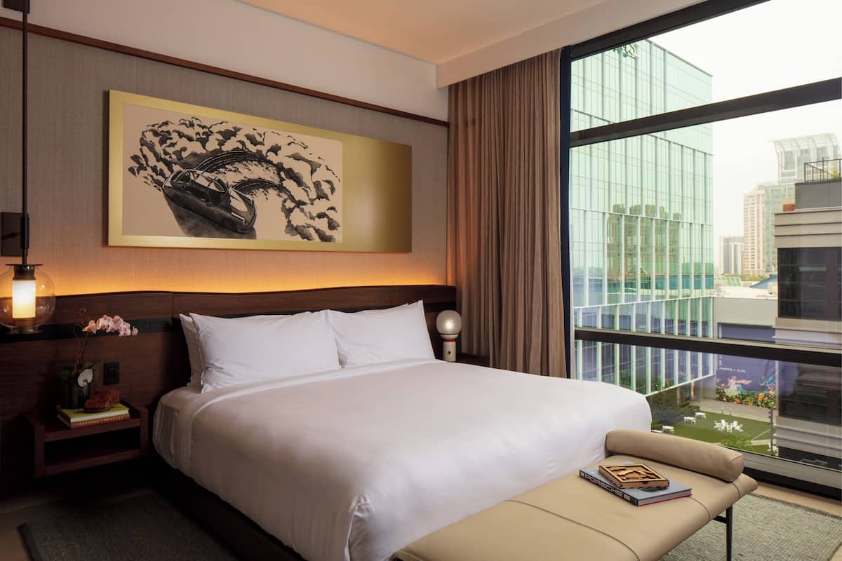 A hotel room with a large bed, a bedside table with a lamp, a bench with a book, a large window with a city view, and a framed artwork on the wall.