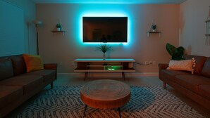 Un salón moderno con un televisor de pantalla plana grande montado en la pared, una mesa de centro de madera, dos sofás y plantas decorativas.