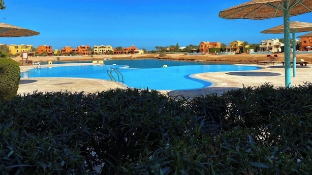 Una piscina con più corsie, circondata da sdraio e ombrelloni, con una fila di edifici colorati sullo sfondo.