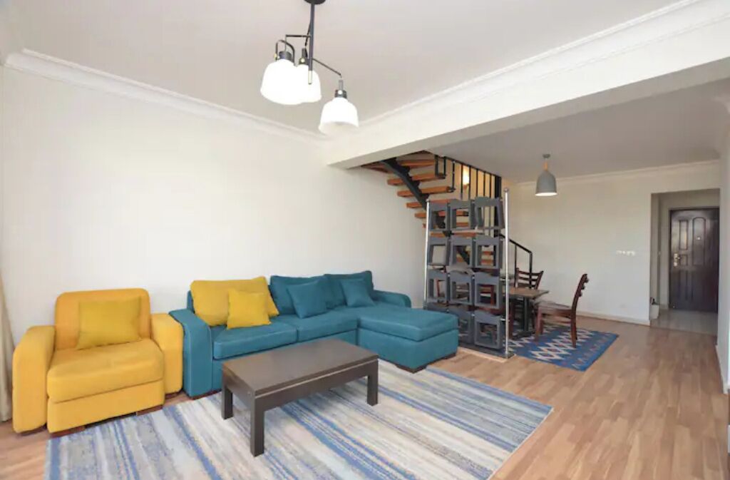A living room with a yellow sofa, a blue sectional sofa, and a coffee table. There is a staircase leading to a loft area with a desk and chair.
