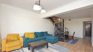 Een woonkamer met een gele bank, een blauwe hoekbank en een salontafel. Er is een trap die leidt naar een vide met een bureau en stoel.