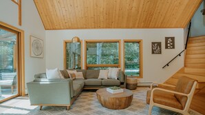 Un salon moderne avec un canapé d’angle, une table basse ronde et une chaise en bois. De grandes fenêtres laissent entrer la lumière naturelle et offrent une vue sur l’extérieur.