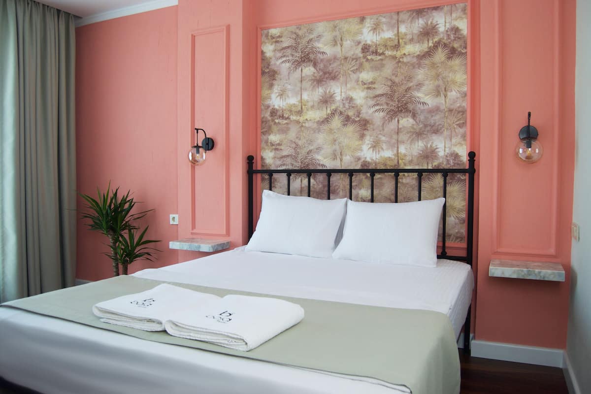 Quarto de hotel com cama, cabeceira estampada com padrão floral, uma pequena planta e mesa de cabeceira com abajur.