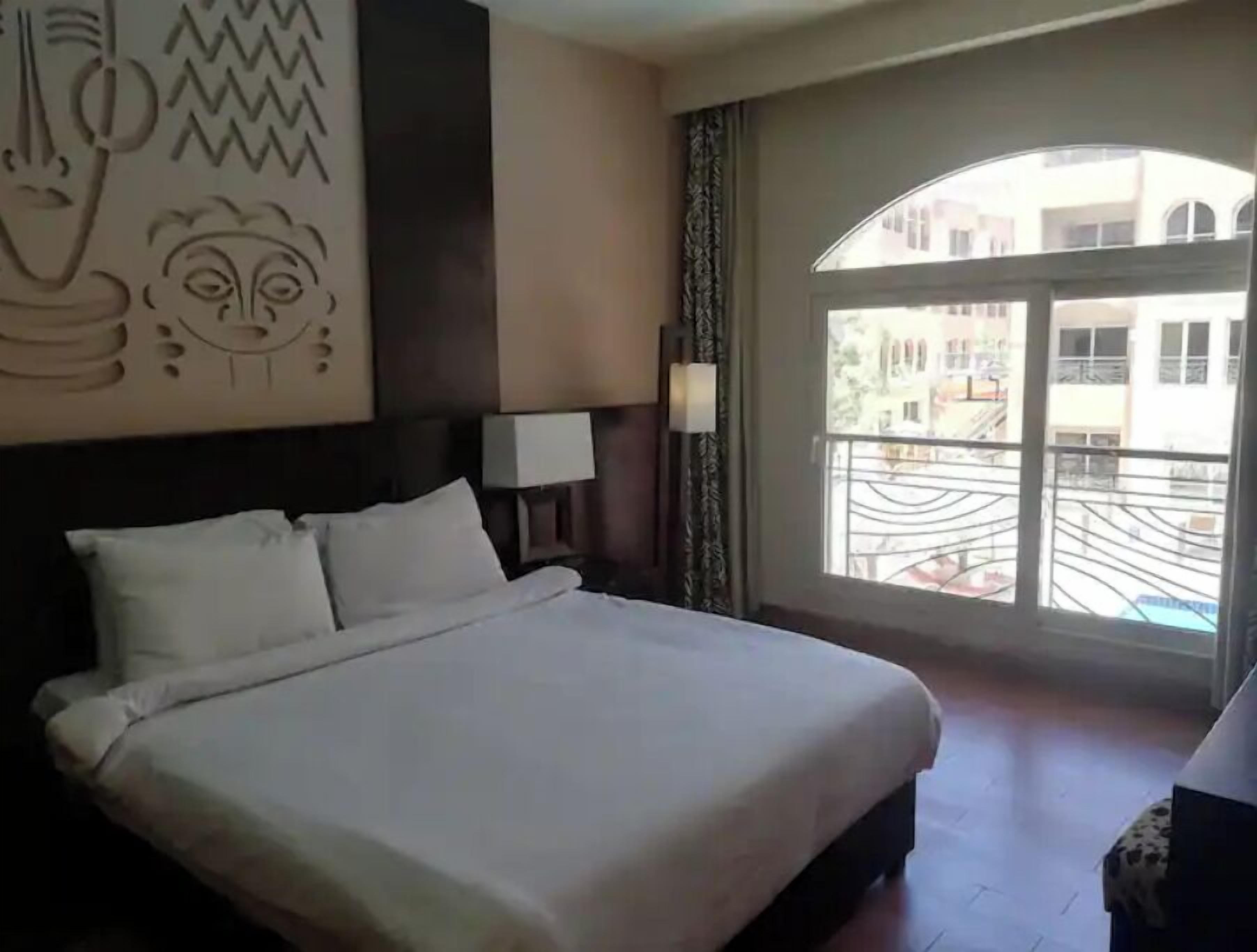 A hotel room with a large bed, a wall-mounted artwork, a window with a view of the outside, and a balcony.
