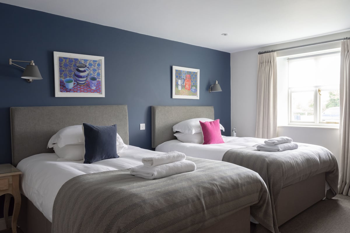 A hotel room with two beds, a nightstand, a lamp, and two framed artworks on the wall.