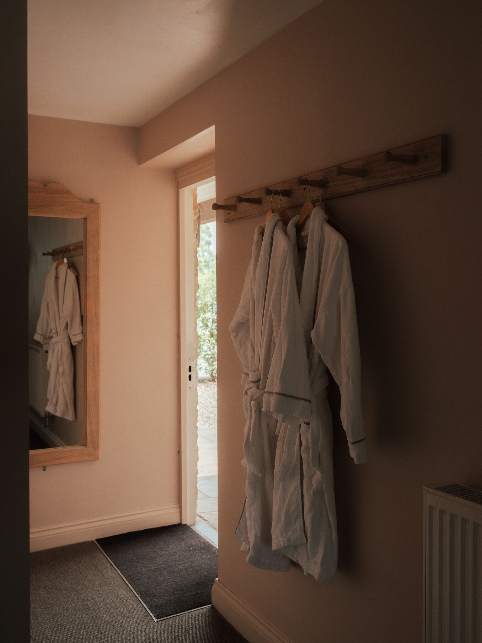 Ein Badezimmer mit einem Holzregal, das weiße Bademäntel, einen Spiegel und eine zur Außenwelt führende Tür beherbergt.