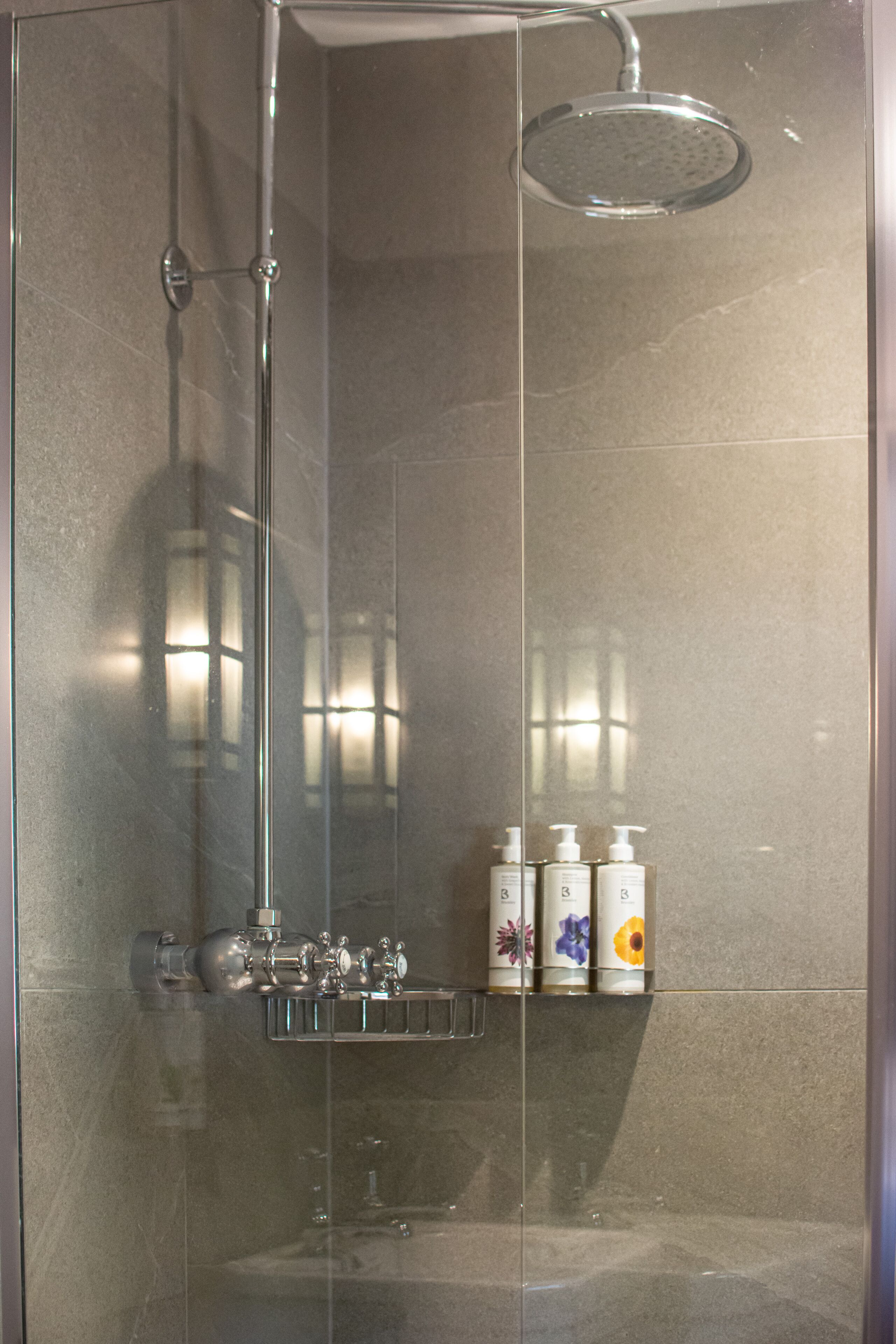 A glass-enclosed shower with a rain showerhead, a chrome faucet, and a shelf with toiletry bottles.
