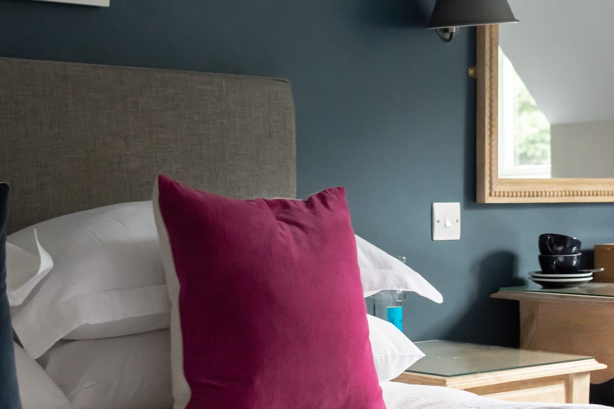 A hotel room with a bed, pillows, a bedside table, a lamp, a mirror, and a painting on the wall.