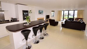 A modern kitchen with a central island, bar stools, a dining area with a sofa, and a large window offering a view of the outdoors.