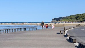 Ein hölzerner Steg führt zu einem Strand, wo Menschen spazieren gehen, im Hintergrund eine felsige Klippe.
