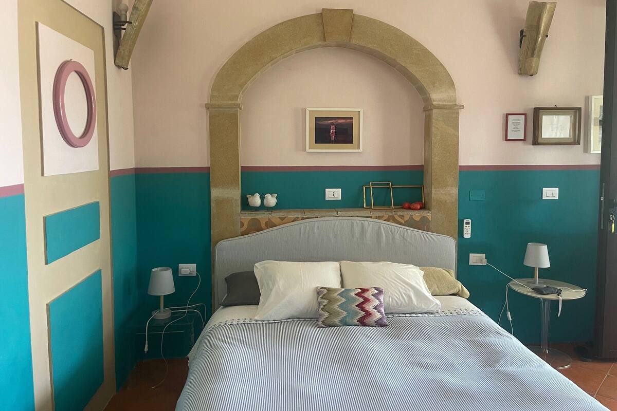 Un dormitorio con cama, una mesita de noche, una lámpara, un cuadro en la pared y una pequeña mesa con lámpara.