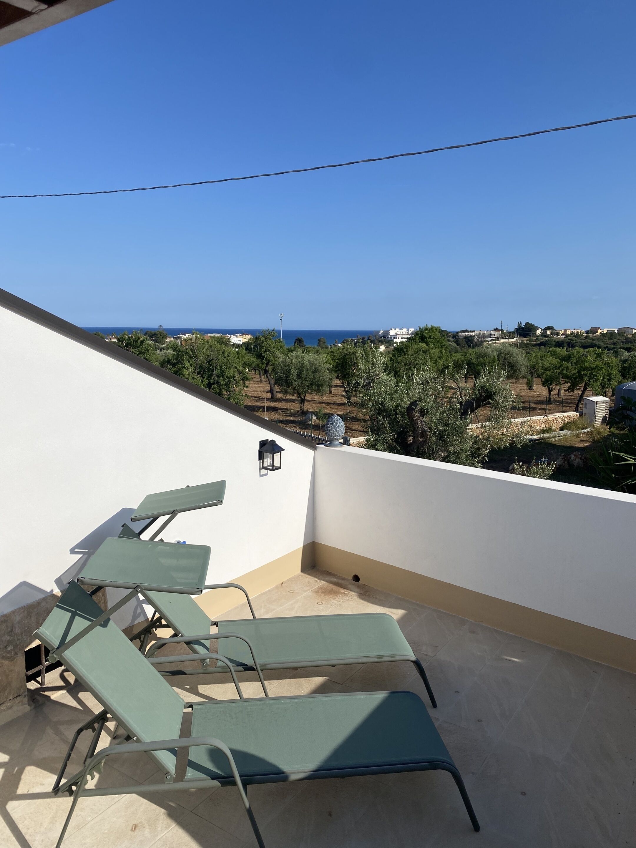 A balcony with green lounge chairs, a wall-mounted lantern, and a view of the sea and distant buildings.