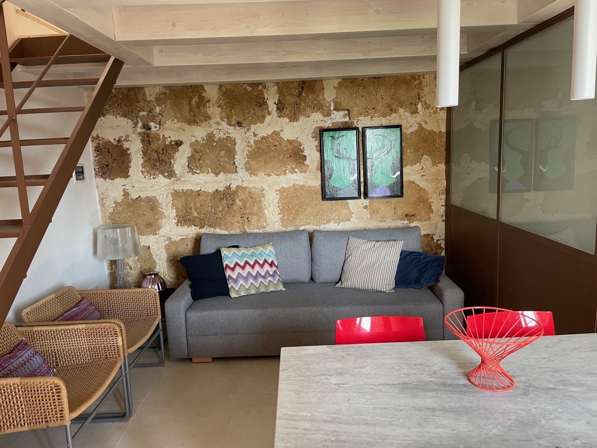 Una sala de estar con una pared de piedra, un sofá gris con cojines estampados, sillas de mimbre y un sillón rojo, junto a una mesa de mármol.