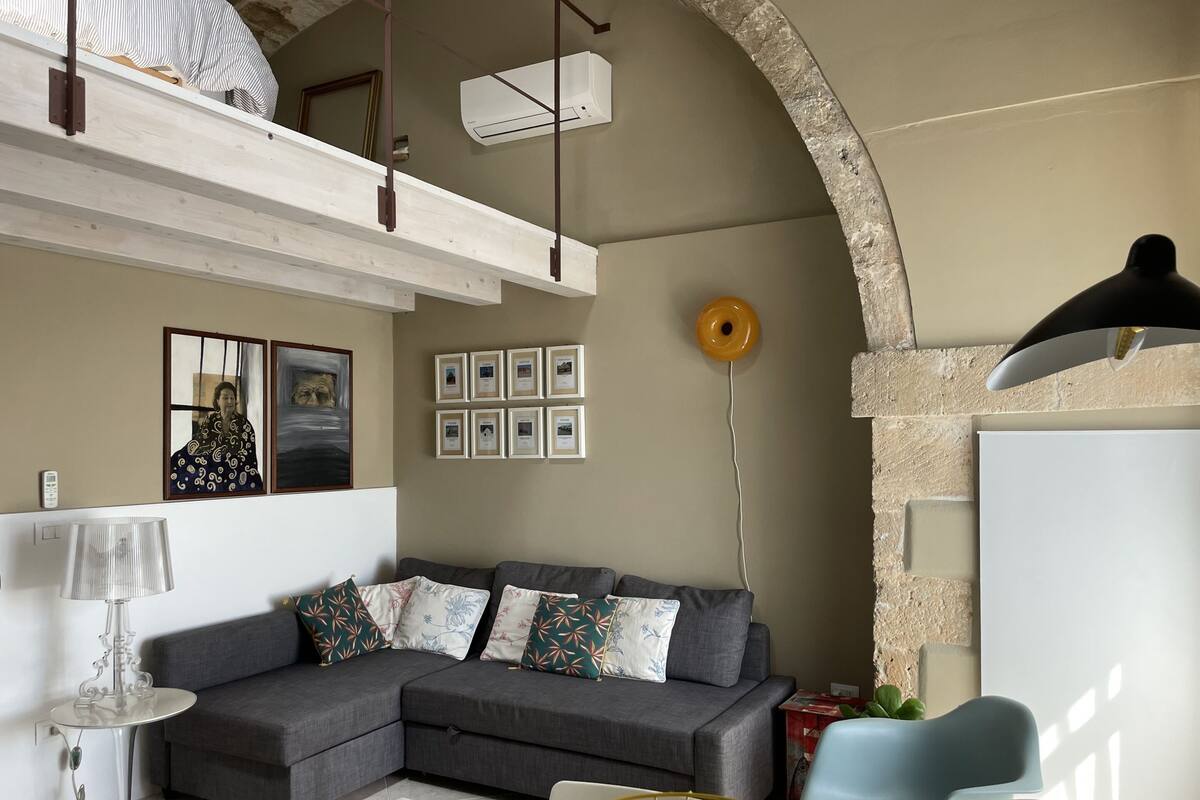 Una sala moderna con un sofá seccional gris, una mesa de centro de vidrio transparente, un sillón azul y una lámpara colgante.