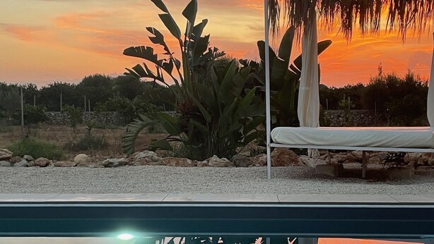 Una piscina con una silla y una estructura de techo de paja, rodeada de vegetación y una vista al atardecer.