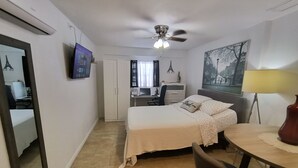 Habitación de hotel con cama, escritorio, silla, ventilador de techo y televisor en la pared.