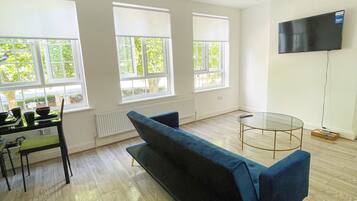 A spacious living room with a blue sofa, a dining table with chairs, a glass coffee table, and a flat-screen TV mounted on the wall.