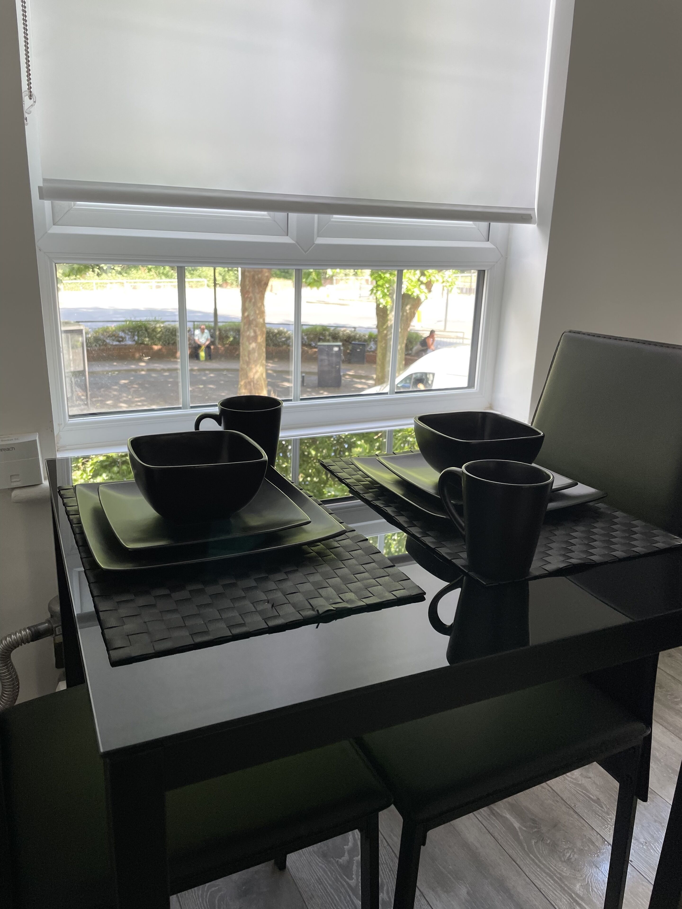 Ein Esstisch mit schwarzen Schüsseln und Tassen, ein Laptop und Blick auf eine Straße und Bäume durch ein Fenster.