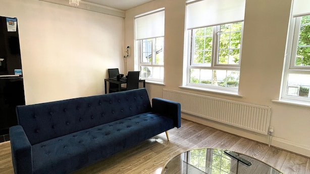 Ein geräumiges Wohnzimmer mit einem blauen Sofa, einem Glastisch und großen Fenstern mit Blick auf Grünflächen.