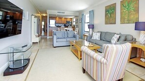 Een moderne woonkamer met een bank, fauteuil, en salontafel, een televisie, en een keuken op de achtergrond.